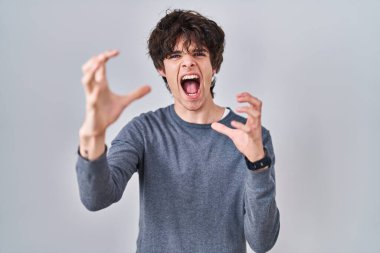 Young man standing over isolated background shouting frustrated with rage, hands trying to strangle, yelling mad 
