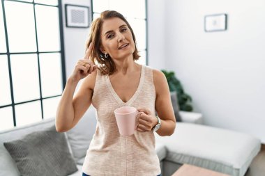 Orta yaşlı bir kadın evde kahve içip gülümsüyor. Tek parmağıyla kafasını işaret ediyor. Harika bir fikir ya da düşünce. 