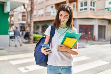 Young beautiful hispanic woman student holding books using smartphone at street