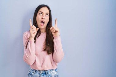 Mavi arka plan üzerinde duran genç esmer kadın şaşırmış ve şaşırmış bir şekilde yukarı bakıyor ve parmaklarıyla işaret ediyor ve kollarını kaldırıyor.. 