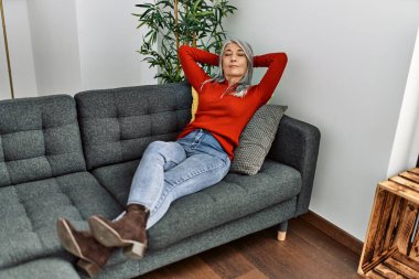 Orta yaşlı, kır saçlı, elleri başının üstünde, evinde kanepede oturan bir kadın.