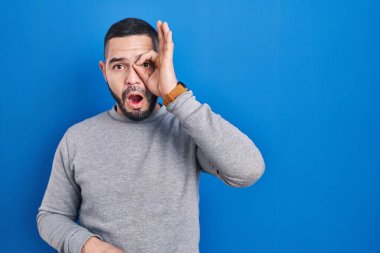 Mavi arka planda dikilen İspanyol adam şaşırmış yüz ifadesiyle şaşırmış, parmaklarından bakan bir hareket yapıyor. İnançsız ifade. 