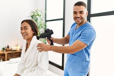 Latin erkek ve kadın fizyoterapi üniforması giyiyorlar boyuna masaj yapıyorlar güzellik merkezinde perküsyon tabancası kullanıyorlar.