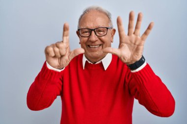 İzole edilmiş arka planda duran yaşlı adam yedinci parmaklarını gösterip gülümserken kendinden emin ve mutlu görünüyor.. 