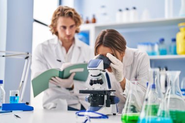 Man and woman wearing scientist uniform writing on notebook using microscope at laboratory