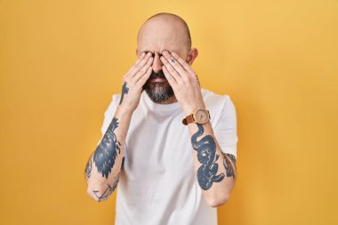 Sarı arka planda dikilen İspanyol dövmeli genç adam yorgunluk ve baş ağrısı için gözlerini ovuşturuyor, uykulu ve yorgun ifade. görme sorunu 