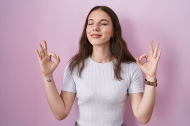 Young hispanic girl standing over pink background relaxed and smiling with eyes closed doing meditation gesture with fingers. yoga concept. 