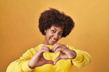 Sarı arka planda dikilen genç Afro-Amerikan kadın. Sevgiyle gülümsüyor. Elleriyle kalp sembolü yapıyor. romantik kavram. 