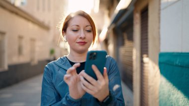 Young redhead woman wearing sportswear listening to music using smartphone at street