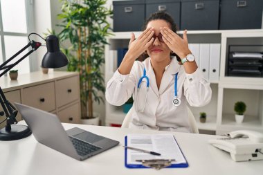 Genç İspanyol kadın doktor üniforması giyiyor ve steteskop ile gözleri kaplıyor neşeli ve komik gülümseyen ellerle. kör kavram. 