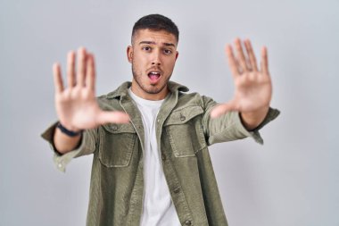 Young hispanic man standing over isolated background doing stop gesture with hands palms, angry and frustration expression 