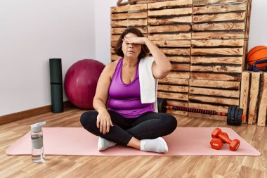 Middle age hispanic woman sitting on training mat at the gym covering eyes with hand, looking serious and sad. sightless, hiding and rejection concept 