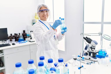 Orta yaşlı, gri saçlı, bilim adamı üniforması giyen bir kadın laboratuarda dizüstü bilgisayar kullanarak test tüpü tutuyor.