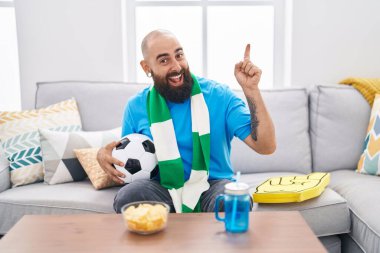 Young hispanic man with beard and tattoos football hooligan holding ball supporting team smiling happy pointing with hand and finger to the side 