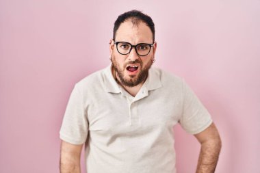 Plus size hispanic man with beard standing over pink background in shock face, looking skeptical and sarcastic, surprised with open mouth 