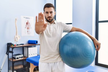 Rehabilitasyon kliniğinde elinde pilates topu tutan yakışıklı adam dur işareti yapıyor ciddi ve kendinden emin bir ifadeyle, savunma jesti. 