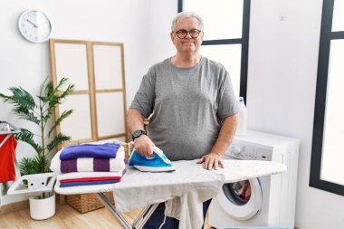 Beyaz üst düzey bir adam evde kıyafetlerini ütülüyor. Olumlu ve mutlu görünüyor. Kendine güvenen bir gülümsemeyle dişlerini gösteriyor. 