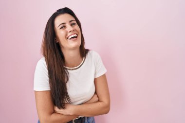 Young brunette woman standing over pink background smiling and laughing hard out loud because funny crazy joke with hands on body. 