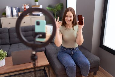 Güzel esmer kadın evde akıllı telefonuyla video kaydı yapıyor. Parmaklarıyla imza atıyor, gülümsüyor. Mükemmel bir sembol. 
