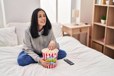 Hispanic woman eating popcorn watching a movie on the bed winking looking at the camera with sexy expression, cheerful and happy face. 