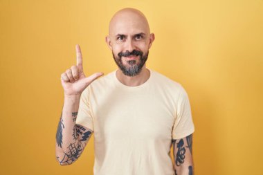 Sarı arka planda dikilen İspanyol dövmeli adam iki numarayı gösterip işaret ederken kendinden emin ve mutlu bir şekilde gülümsüyor.. 