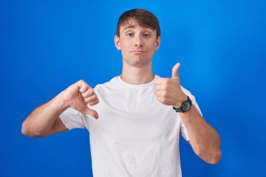 Caucasian blond man standing over blue background doing thumbs up and down, disagreement and agreement expression. crazy conflict 