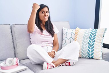 Young hispanic woman sitting on the sofa at home angry and mad raising fist frustrated and furious while shouting with anger. rage and aggressive concept. 