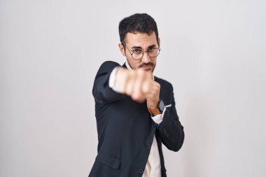 Handsome business hispanic man standing over white background punching fist to fight, aggressive and angry attack, threat and violence 