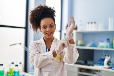 Young african american woman wearing scientist uniform and gloves at laboratory