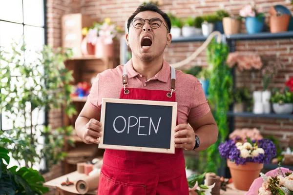 Chinese young man working at florist holding open sign angry and mad screaming frustrated and furious, shouting with anger looking up.
