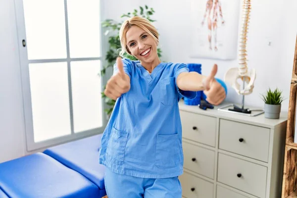 Beautiful blonde physiotherapist woman working at pain recovery clinic approving doing positive gesture with hand, thumbs up smiling and happy for success. winner gesture.