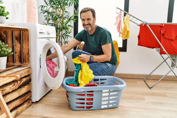髭を生やした中年男性が洗濯機に洗濯機を入れてポジティブで幸せな立ち上がり 歯を示す自信に満ちた笑顔で笑顔 — ストック写真