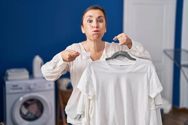 中年时 惊慌失措的女人拿着衬衫挂在衣架上 脸上挂着洗衣粉 脸上带着滑稽的表情 嘴被空气吹胀 — 图库照片