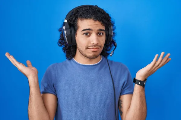 ヘッドフォンを使用して音楽に耳を傾け巻き毛を持つヒスパニック系の男性は 腕や手を上げと混乱表現 疑わしい概念 — ストック写真
