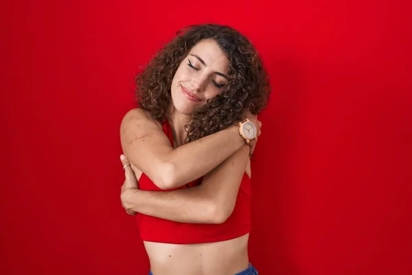 Kıvırcık Saçlı Spanyol Kadın Kırmızı Arka Planda Durup Kendine Sarılıyor — Stok fotoğraf
