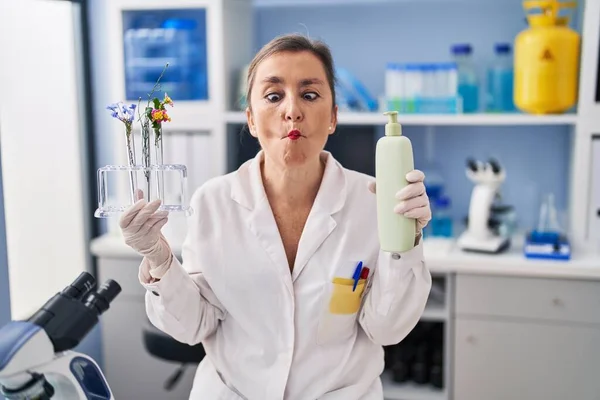 Middle age hispanic woman working at cosmetics laboratory making fish face with mouth and squinting eyes, crazy and comical.
