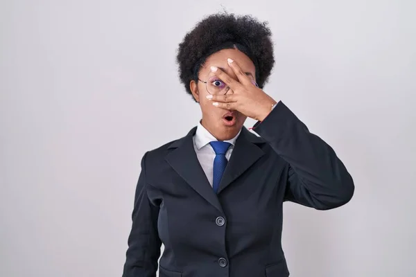 Beautiful african woman with curly hair wearing business jacket and glasses peeking in shock covering face and eyes with hand, looking through fingers with embarrassed expression.