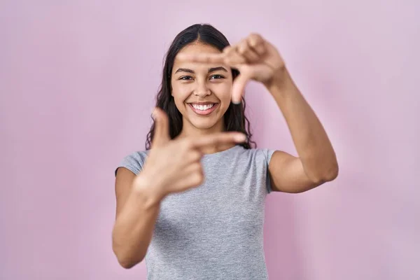 Young brazilian woman wearing casual t shirt over pink background smiling making frame with hands and fingers with happy face. creativity and photography concept.