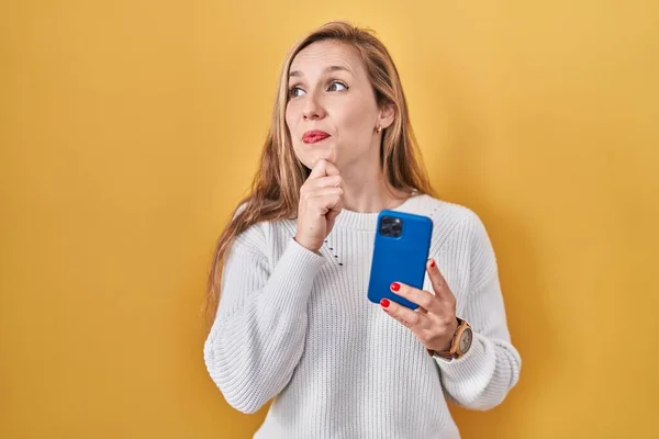 質問について考える顎の手でスマートフォンの入力メッセージを使用して若いブロンドの女性 集中的な表現 笑顔で思いやりのある顔 疑わしい概念 — ストック写真