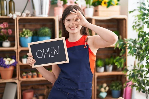 Young caucasian woman working at florist holding open sign smiling happy doing ok sign with hand on eye looking through fingers