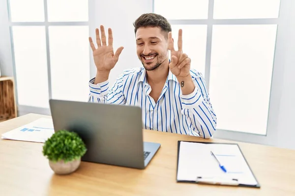 自信と幸せ笑顔ながら コンピュータのラップトップを使用してオフィスで働くひげを持つ若いハンサムな男は指の番号7で示すと指摘します — ストック写真