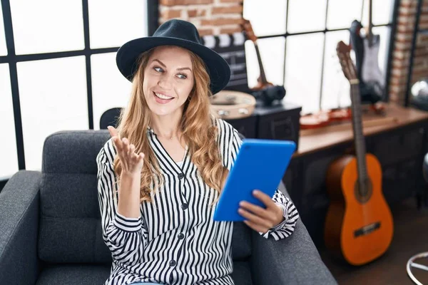 Beautiful blonde woman at music studio using tablet celebrating achievement with happy smile and winner expression with raised hand