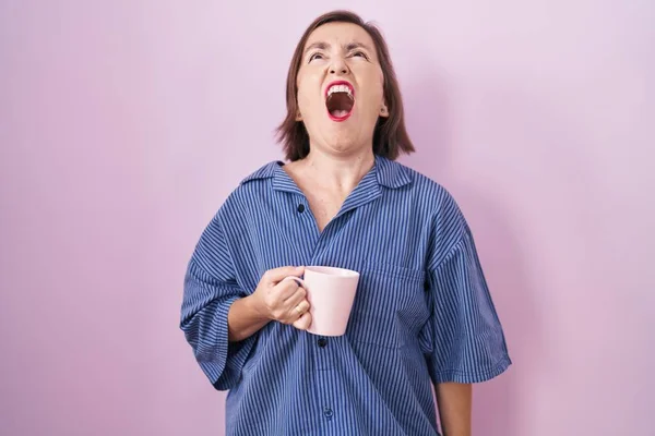 Middle age hispanic woman drinking a cup coffee angry and mad screaming frustrated and furious, shouting with anger. rage and aggressive concept.