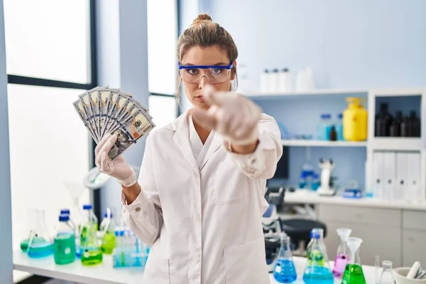 Young woman working at scientist laboratory holding money pointing with finger to the camera and to you, confident gesture looking serious