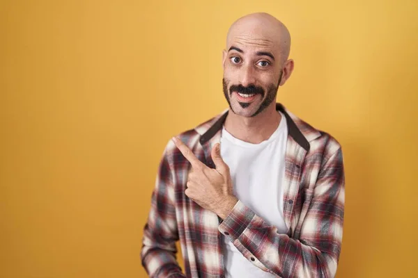 Hispanic man with beard standing over yellow background cheerful with a smile of face pointing with hand and finger up to the side with happy and natural expression on face