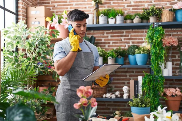 Young hispanic man florist talking on smartphone reading clipboard at flower shop
