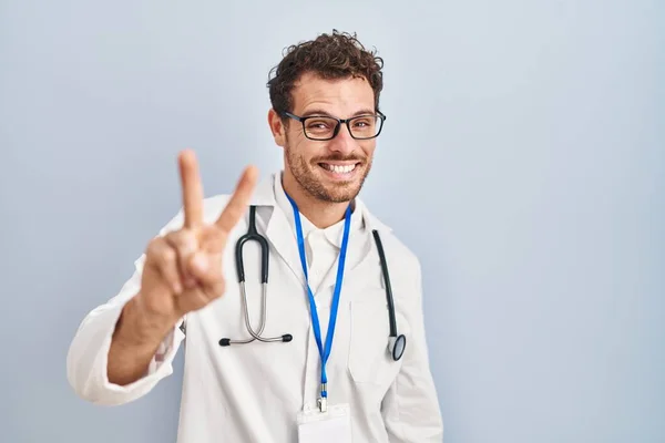 身穿医生制服和听诊器的年轻人微笑着看着摄像机 手指头在做胜利的标志 第二点 — 图库照片