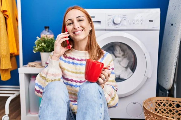年轻的高加索女人在洗衣房用智能手机喝咖啡等洗衣机 — 图库照片