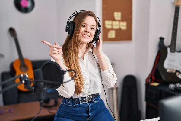 Young caucasian woman recording song at music studio smiling happy pointing with hand and finger to the side