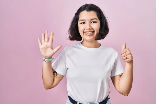 Young hispanic woman wearing casual white t shirt over pink background showing and pointing up with fingers number six while smiling confident and happy.